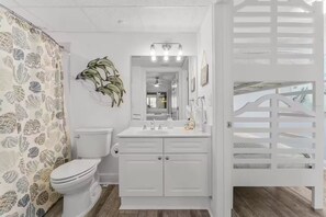 A bathroom with a toilet, sink, and mirror. There is a plant on the wall and a shower curtain with a shell pattern. - Sunset Views, Beachfront & Resort Pools (Panama City Beach)