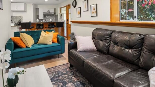 A modern living room with a teal sofa, a leather couch, and a coffee table. There is a kitchen in the background with white cabinets and a window with pink flowers.