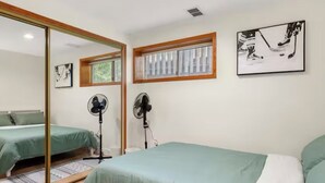 A bedroom with two beds, a large mirror, two standing fans, and a window with blinds.