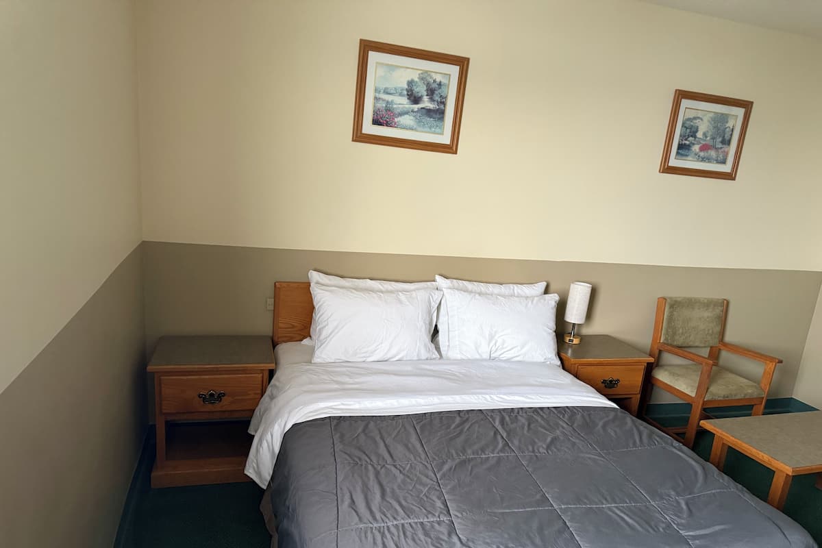 A neatly made bed with white linens, a wooden nightstand with a lamp, and a chair.