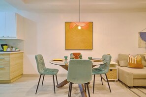 A modern dining area with a round glass table, two chairs, a hanging light fixture, and a sofa with cushions.