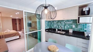 A modern hotel room with a glass door leading to a balcony, a kitchenette with a sink and stove, a dining table with a wooden board, and a unique spherical light fixture. - Serrambi Beach Flat - Porto de Galinhas (Ipojuca)