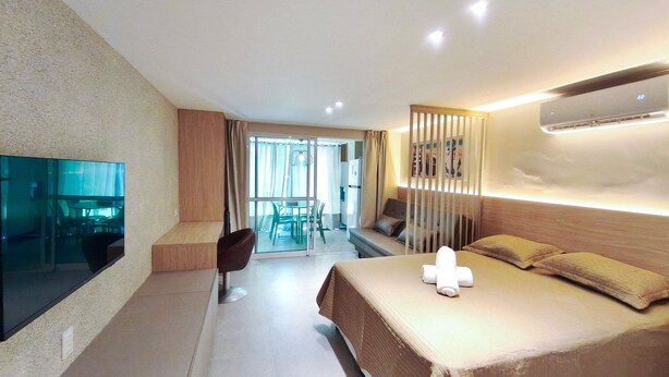 A modern hotel room with a bed, a TV, a seating area with a table and chairs, and a view through a glass door to the outside. - Serrambi Beach Flat - Porto de Galinhas (Ipojuca)