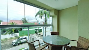 A balcony with wicker chairs and a glass table, overlooking a street with parked cars and buildings.