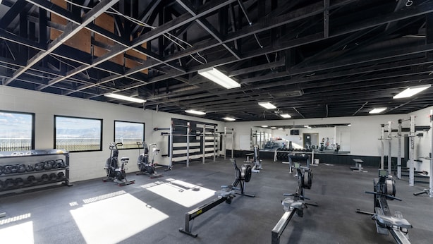 A gym with various exercise equipment, large windows, and a high ceiling with exposed beams. - Utah Prep Basecamp (Hurricane)