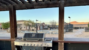 An outdoor barbecue grill under a wooden pergola with a view of a fenced sports court and buildings in the background. - Utah Prep Basecamp (Hurricane)