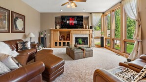 A spacious living room with a fireplace, leather sofas, a glass coffee table, and large windows offering a view of the outdoors.
