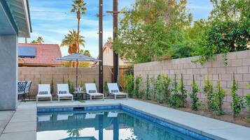 A backyard with a swimming pool, white lounge chairs, and a brick wall.