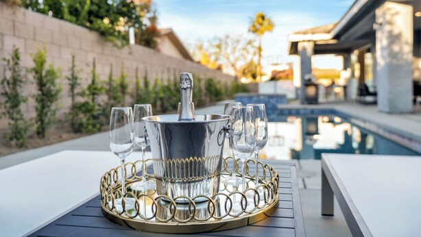 Mesa junto a la piscina con un cubo de champán y copas, todo sobre una bandeja decorativa.