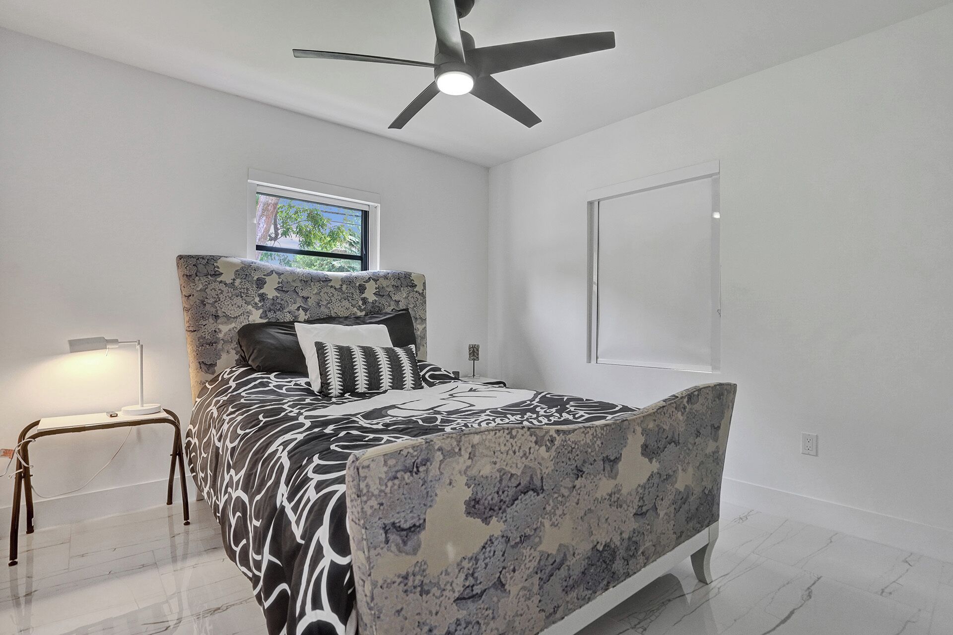 A bedroom with a bed, a ceiling fan, a nightstand, a lamp, and a window with a view of trees.