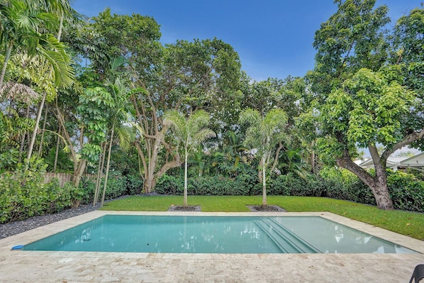 A rectangular swimming pool with a tiled deck, surrounded by lush greenery and trees.