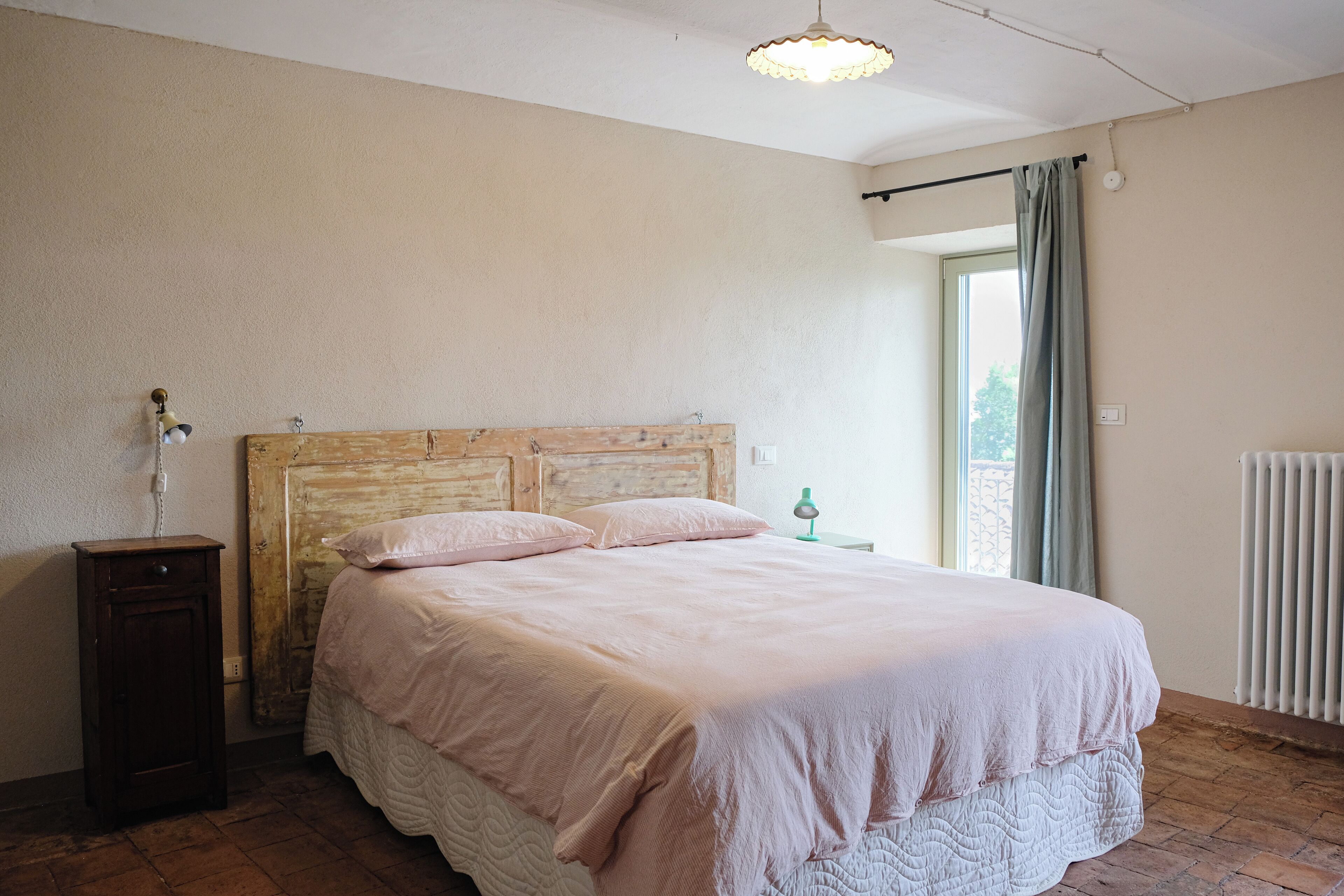 Un dormitorio con cabecera de madera, cama con ropa de cama rosa, mesita de noche, lámpara y ventana con cortinas.