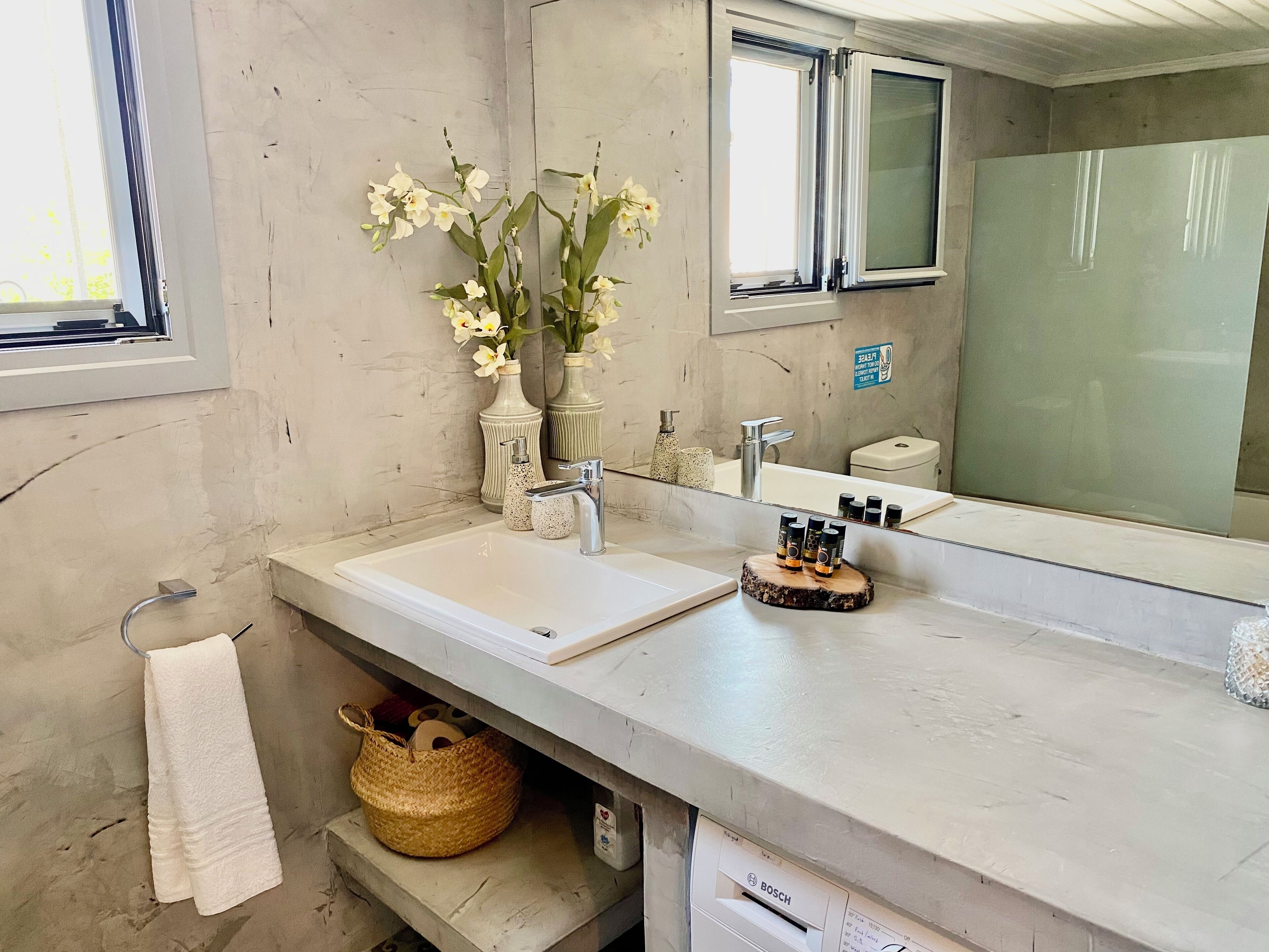 A bathroom with two sinks, a mirror, and a window. There are vases with flowers, a towel rack, and a basket.