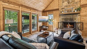Een gezellig cabin interieur met een stenen open haard, leren banken, een televisie en grote ramen met uitzicht op een bebost gebied.