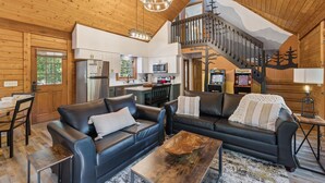 Een ruime woonkamer met twee zwarte leren banken, een houten salontafel en een eettafel met stoelen. In de achtergrond is een keuken met moderne apparatuur zichtbaar.