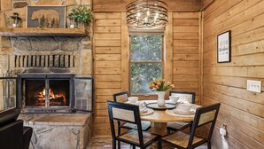 Een gezellig chaletinterieur met een stenen open haard, houten wanden, een eettafel met stoelen en een raam met uitzicht op bomen.