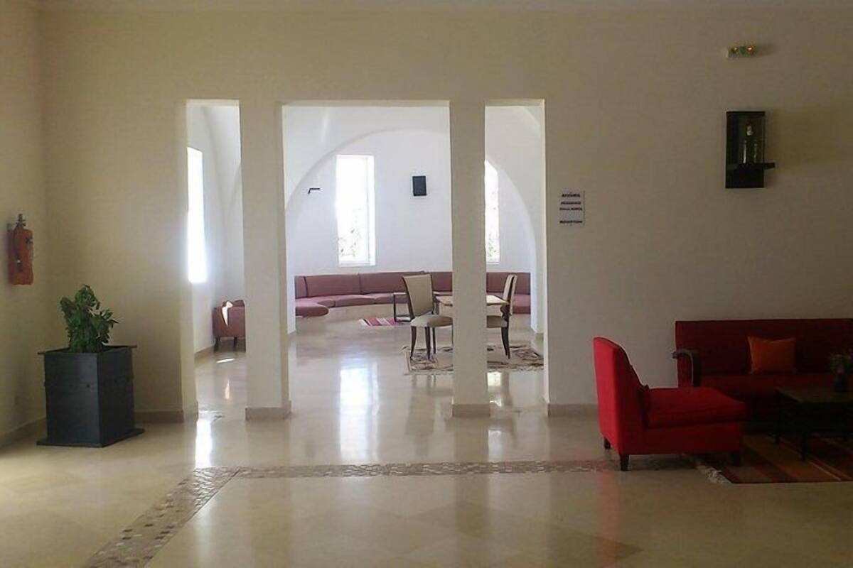 Un hall d’entrée spacieux, avec un canapé rouge, un pot de fleurs noir et une grande porte en arc donnant sur un espace détente.