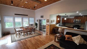 A spacious living room with wooden flooring, a dining area with a table and chairs, a kitchen with wooden cabinets and a microwave, and a large window with blinds. - Spacious, modern, home by the lake! (Northville)