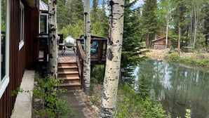 A wooden deck with a table and chairs, a small bridge leading to a cabin, and a river in a forested area.