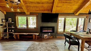 A cozy cabin interior with a wooden ceiling, a fireplace, a TV mounted on the wall, and a dining area with a bench and chairs.