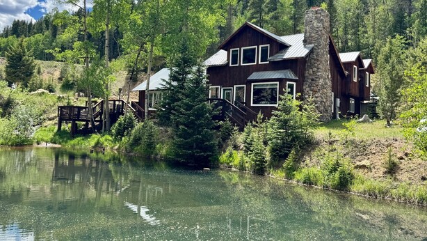 A cabin with a stone chimney, wooden deck, and a view of a lake surrounded by trees.