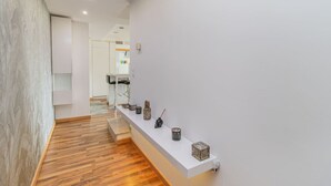 Um corredor moderno com piso de madeira, uma parede branca com uma prateleira exibindo objetos decorativos e um vislumbre de uma área de cozinha ao fundo.