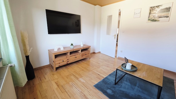 Una habitación con piso de madera, un televisor montado en la pared, un mueble de madera con cajones, una pequeña planta y una puerta que da a otra habitación.