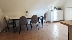 Un comedor con una mesa blanca, cuatro sillas grises, un jarrón pequeño con flores secas y un armario blanco con cajones.