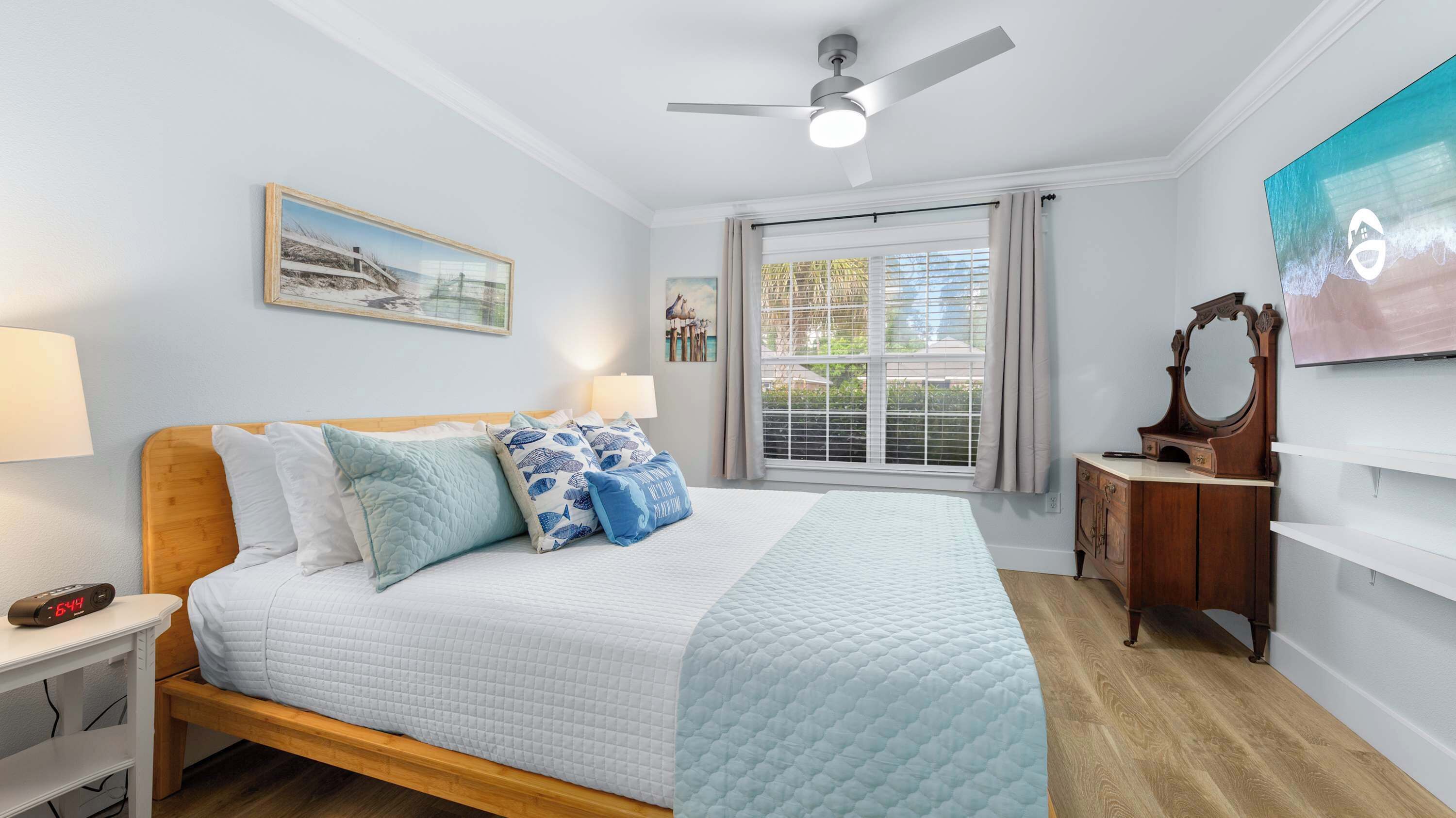 A bedroom with a bed, bedside lamps, a ceiling fan, a window with blinds, a painting, and a TV.