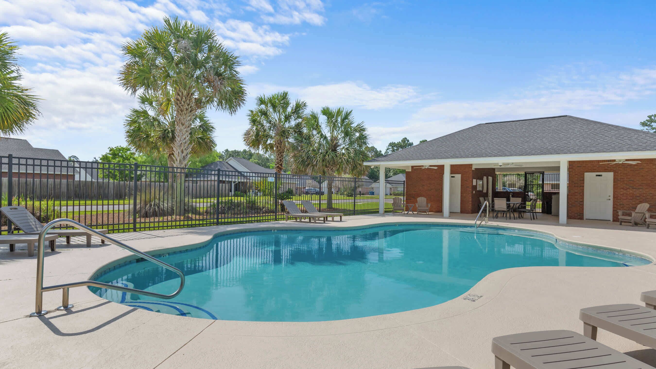 A swimming pool with lounge chairs, a covered patio area, and a fenced yard with palm trees.