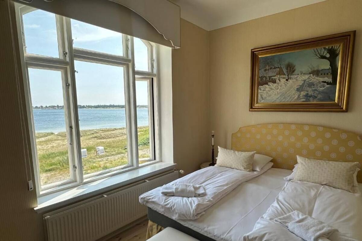 Un dormitorio con un ventanal grande que da a una masa de agua, una cama con sábanas blancas, un cabecero amarillo con lunares y un cuadro enmarcado en la pared.
