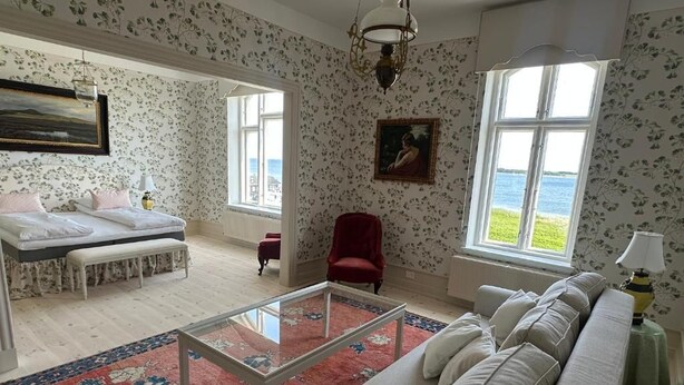 A spacious living room with a large sofa, a glass coffee table, and a view of the sea through large windows. - Aaroesund Badehotel (Haderslev)