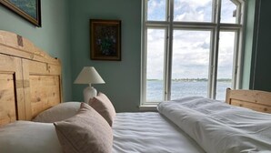 A bedroom with a wooden headboard, a bedside lamp, and a view of the water through a large window. - Aaroesund Badehotel (Haderslev)