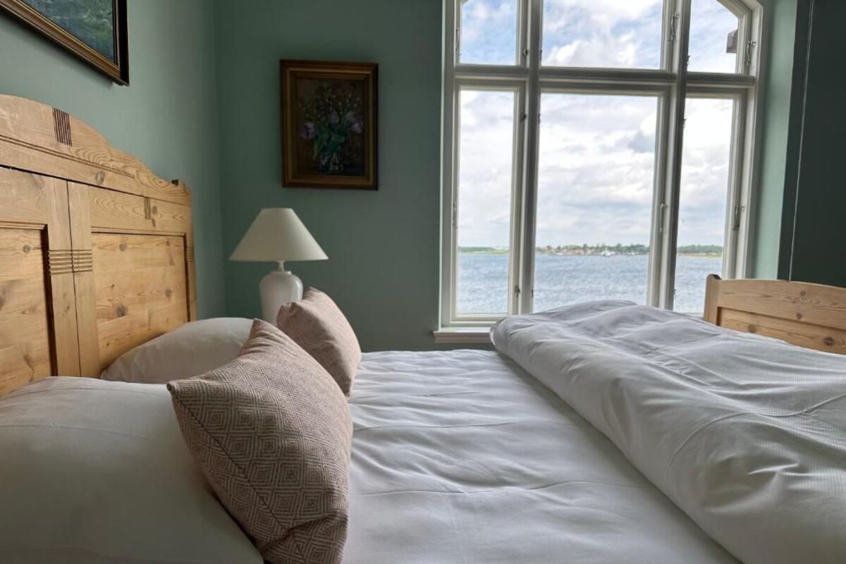A bedroom with a wooden headboard, a bedside lamp, and a view of the water through a large window.