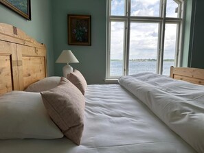 A bedroom with a wooden headboard, a bedside lamp, and a view of the water through a large window. - Aaroesund Badehotel (Haderslev)