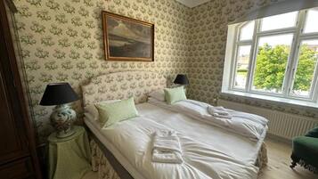A bedroom with two beds, floral wallpaper, a painting above the beds, a window with a view of greenery, and a green chair.