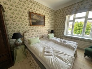 A bedroom with two beds, floral wallpaper, a painting above the beds, a window with a view of greenery, and a green chair. - Aaroesund Badehotel (Haderslev)