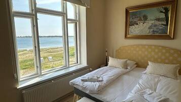 A bedroom with a large window overlooking a body of water, a bed with white linens, a yellow polka-dotted headboard, and a framed painting on the wall.