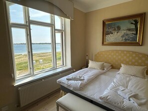 A bedroom with a large window overlooking a body of water, a bed with white linens, a yellow polka-dotted headboard, and a framed painting on the wall. - Aaroesund Badehotel (Haderslev)