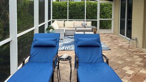Eine überdachte Terrasse mit zwei Liegestühlen und einem Tisch, einer Glaseinfassung und Blick auf ein Haus und Grünflächen.