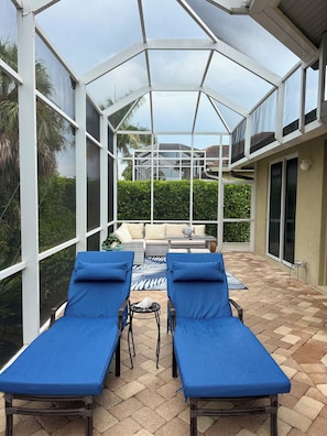 A covered patio with two lounge chairs and a table, a glass enclosure, and a view of a house and greenery.