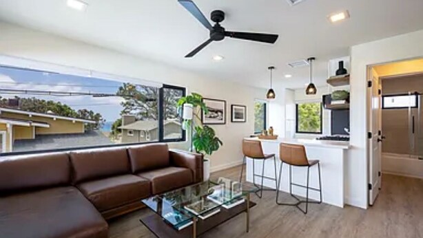 A modern living room with a brown leather sofa, a glass coffee table, and a ceiling fan. - Charming Ocean View 2-bedroom apartment with AC in Del Mar! (Del Mar)