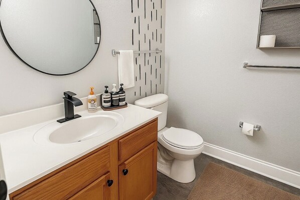 A modern bathroom with a round mirror, a white sink, wooden cabinets, a toilet, and a patterned wall. - 3BR Townhome w/ Pool & BBQ Near Disney (Kissimmee)