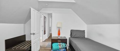 A small, narrow room with a sloped ceiling, a bed, a bedside table with a lamp, and a bench with storage underneath.