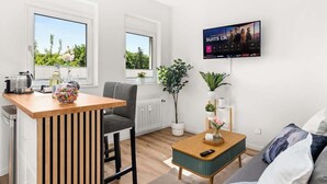 A modern living room with a kitchen area, a TV mounted on the wall, and large windows offering a view of greenery. - MOLA Apartments-Style & Design Wetter Ruhrgebiet (Wetter)