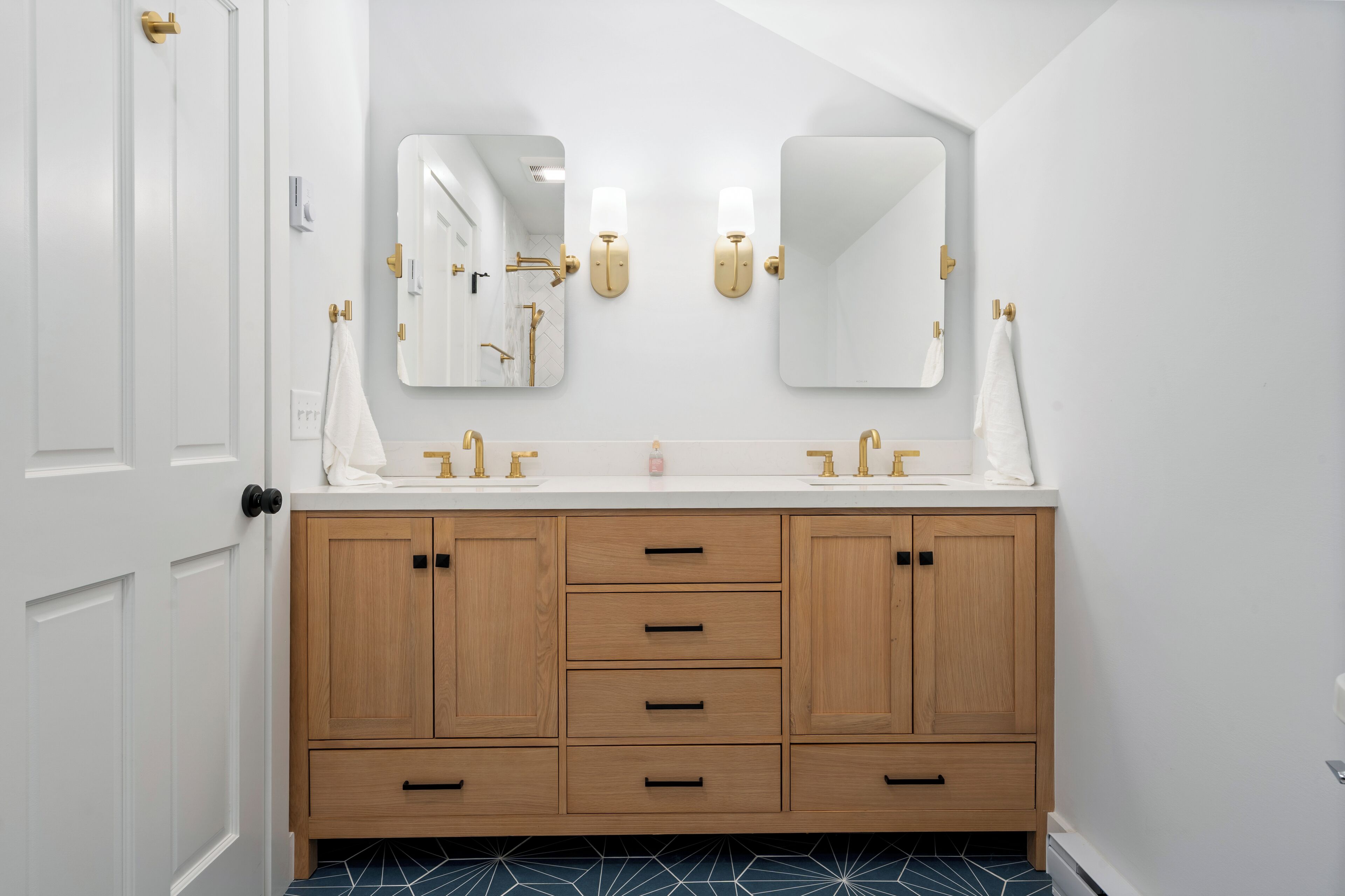 Een badkamer met een witte deur, een houten wastafelmeubel met een dubbele wasbak en twee spiegels aan de muur.