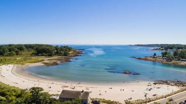 Een kuststrand met helder water, een zandstrand en enkele gebouwen op de achtergrond.