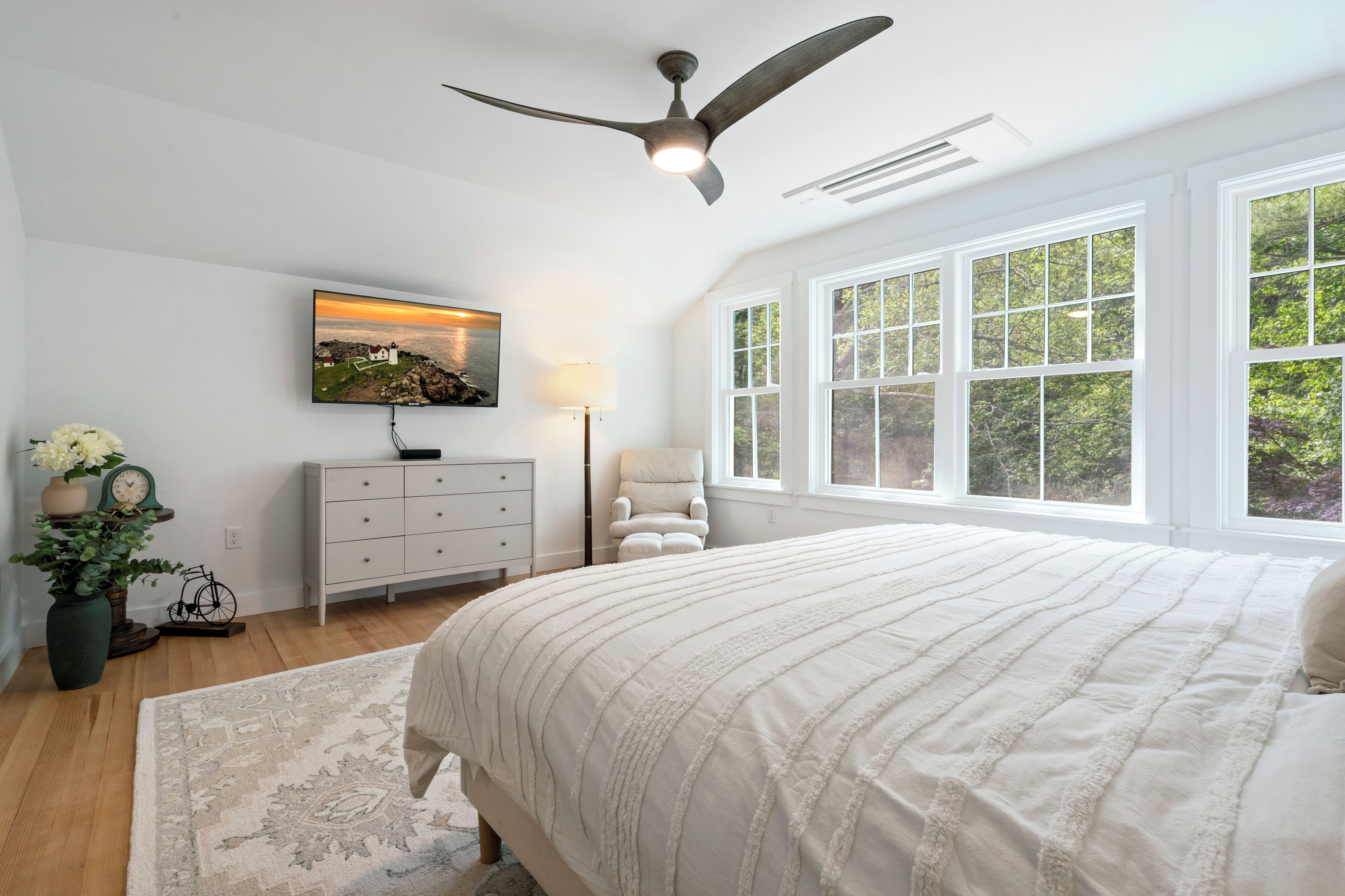 A bedroom with a large bed, a ceiling fan, a TV mounted on the wall, a window with a view of greenery, and a bedside table with a lamp.