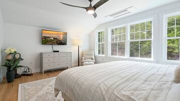 A bedroom with a large bed, a ceiling fan, a TV mounted on the wall, a window with a view of greenery, and a bedside table with a lamp.