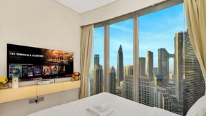 A hotel room with a large bed, a flat-screen TV displaying 'The Umbrella Academy' series, a city view through large windows, and a small table with a vase and a lamp.